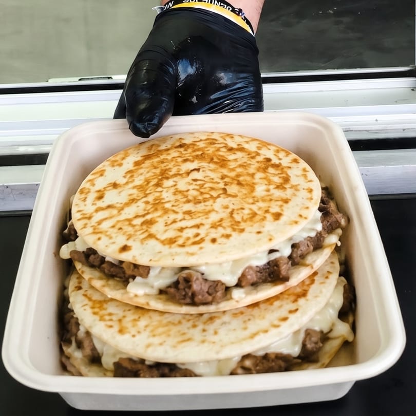 Gloved hand presenting a takeout container filled with stacked quesadillas, featuring meat and melted cheese.