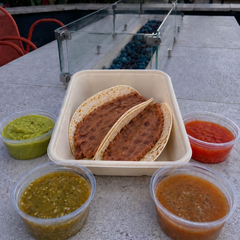 Two bean tacos in a takeout container with four sides of salsa and guacamole on a patio table.